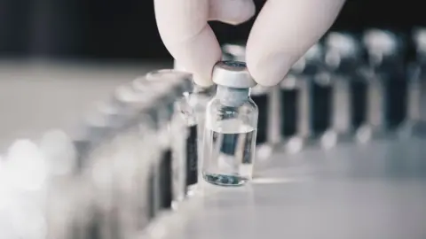 A hand in a white latex glove picks up a single vial between thumb and forefinger. The glass vial contains a clear fluid and has a sliver lid. It is one of a long row of vials disappearing out of focus.