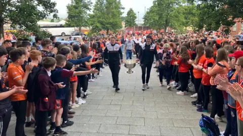 Two men holding either side of the Sam Maguire cup, carrying it though a group of children who are lining their path and clapping