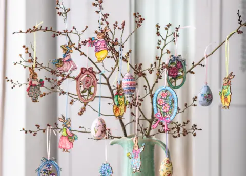 Amy Swann A tree cutting with small buds on it in a green vase with Easter decorations made by Amy hanging from the branches. The decorations are woodcuts of anthropomorphised rabbits in colourful clothing, eggs, and bunches of flowers. They are all colourful with pink, light blues, yellow and light green featuring in the designs.