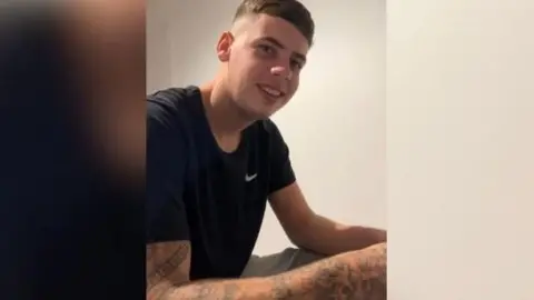 Avon and Somerset Police Wearing a black Nike t-shirt, Tyler smiles at the camera. He is sat down, and tattoos can be seen on his right arm.