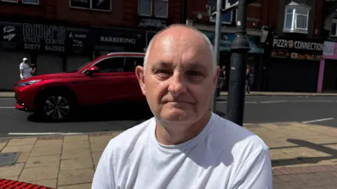 Gemma Dillon A man wearing a white t shirt is sat on a bench. A main road is in the background with a red car. 