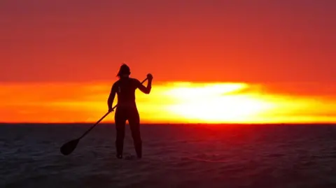 Owen Humphreys/PA Wire View of a paddleboarder silhouetted against a sunrise at sea. The sky is a fiery orange and yellow, and the sea is dark.