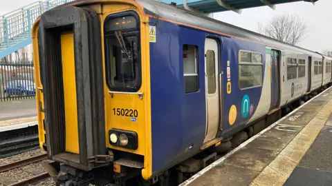 A passenger train, number 150220, on the railway line at Brigg station. The train has blue and grey sides, with "Northern" logos, and a yellow front with glass windows. Grey platforms can be seen, along with a passenger bridge with light blue and white railings.