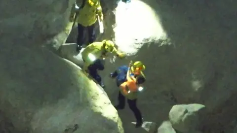 Several people in yellow hi-vis jackets and hard hats walk down boulders with one carrying a boy in an orange shirt.