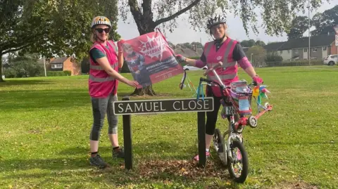 A street sign saying "Samuel Close" with a woman on the left of the sign, and on the right is Rachel Liew who is on a scooter. They are both wearing a helmet and pink hi vis jacket, and holding a sign saying "Sam's Superheroes"