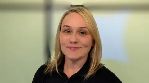 Anna Webb Anna Webb smiling. She has blonde hair and is wearing a black polo shirt. The background has a blur filter effect in place.