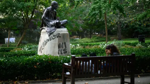  Words and slogans including "Gandhi - Modi Hindu-stani Terrorists" and "Terrorist" are seen on a statue of Mahatma Gandhi inside a park as three people on a bench sit in the foreground