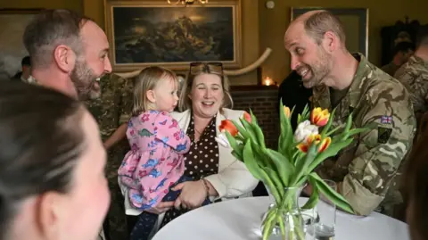 PA Media The Prince of Wales pictured smiling at a baby being held by a woman. Another man can be seen to the left of the image, and also a woman by the camera. There is a bunch of flowers in a vase on the table.