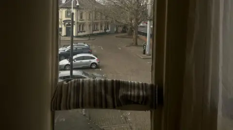 A narrow window with the view to a car park. There is a cream building and trees with no leaves. There are four cars parked. The window has a towel across the middle that is being used to block draughts.