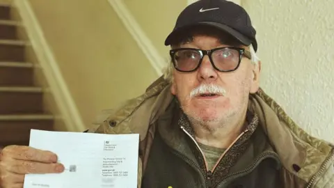 LDRS Arthur Bowling is wearing a black baseball cap, glasses, a green top, two thicker tops and a coat, while holding a letter from the Department for Work and Pensions