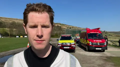 A man with short brow hair looking ahead. He is wearing a black top with a light grey jacket on top. In the background is a red van with 'Fire' written on the front and a red and yellow 4x4 vehicle.