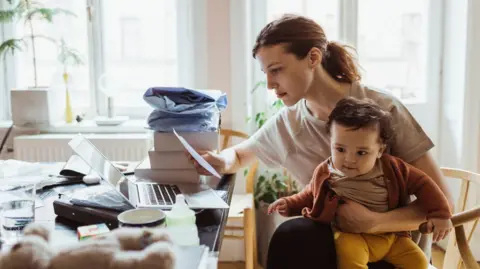 A woman holding her infant son on her lap while reading bills at her desk.