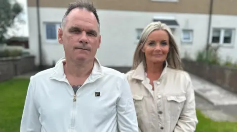 Veteran Craig Kirkland, a man with short grey hair, wears a while zipper top and stands beside his wife Caroline, a blonde woman in a cream denim jacket.