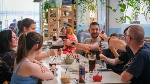 A general view of the cafe. In the foreground are six people sitting around a table. A few of them are pointing plastic guns at each other. There are cards set out on the table. Everyone looks amused.