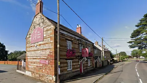 Google The exterior of the White Hart pub, which is a stone-built two-storey building with small porch over the door. It has big red signs on the side and above the door advertising the pub. There are two big hanging baskets with pink flowers underneath the upper floor windows. It is next to a road and there is a car park behind.