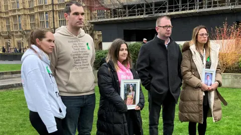 Five people standing outside the Houses of Parliament. Some of them are holding pictures of loved ones.