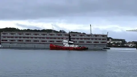 A grey and red three-storey barge being towed by a red and white tug boat in open water. 