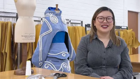 A woman with glasses and long dark hair smiles, wearing a dark blouse, at the camera. She is sitting next to a desk with scissors and a measuring tape with a dress on a mannequin behind her.