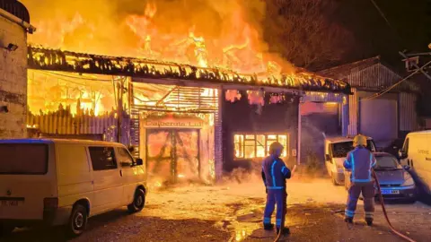 A burning industrial unit with flames coming through the roof. There are three vans and a car parked outside. Two firefighters are holding hoses with steam and smoke around them. 