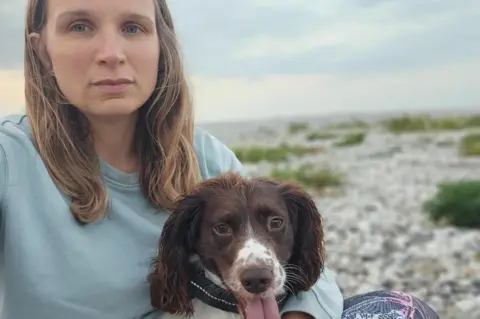 Laura Timmis Laura, who was long blonde hair, wearing a blue sweatshirt, and holding a brown and white spaniel. They are sat on a pebbly beach with the sky and sea in the background. 