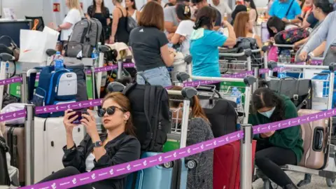 2024年7月19日、香港国際空港のチェックイン列で手荷物カートに座るサングラスをかけた女性。 