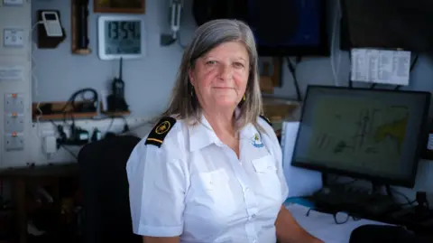 Photo provided by the South West Coast Path Association, Photographer: Keith Mason A woman in a white shirt is sitting at a computer desk and is looking into the camera. Technology and radio equipment is visible behind her. 