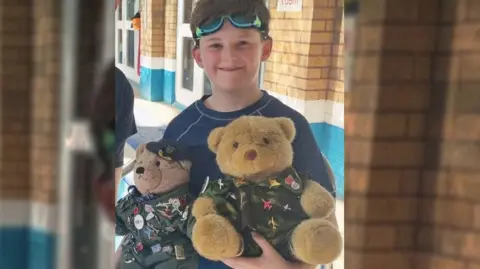 A child stands in his goggles beside a pool holding two mascot teddy bears.