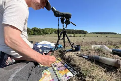 SHAUN WHITMORE/BBC Darren Woodhead, wearing a white T-shirt and grey cap mixes watercolours sitting on the floor and painting the landscape around him.