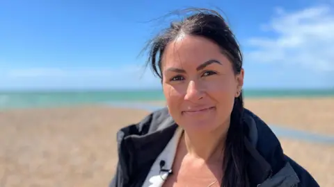 Juliette Parkin/BBC A dark-haired woman, wearing a white shirt and black coat. She is standing at a pebbled beach. 