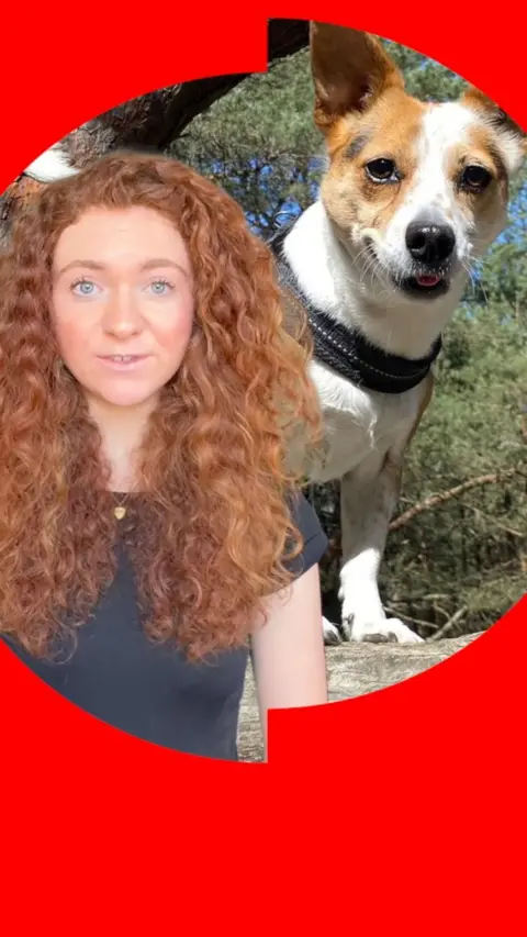 A woman with red curly hair stands in front of a picture of a dog in the woods