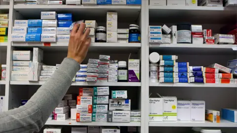 An arm with a grey sleeve is reaching out to a set og shelves packed with a variety of medications