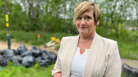 Candy Manders has short honey blonde hair and is wearing a beige blazer and white top. She is standing in front of green grass which has a pile of black binbags on it.