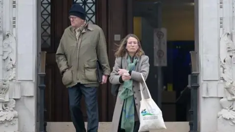 A woman and a man coming down steps outside Newport Crown Court. The man, on the left, wears dark blue trousers and a khaki coat, and a dark blue woolly hat. The woman, on the righ, wears a grey coat and green patterned scarf; she carries a tote bag. 
