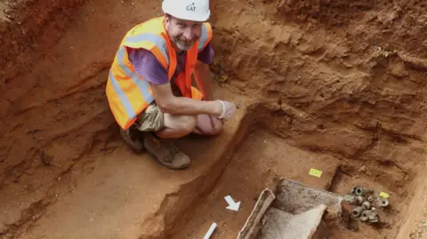 Colchester Archaeological Trust Archaeologist Alec Wade wearing a hi-vis vest and white helmet while crouching next to the coffin, which is surrounded by dried mud.