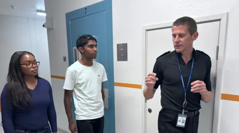 BBC/Kate Bradbrook A young woman (left) and young man (middle) in a blue and white corridor speaking to police chief Ivan Balhatchet (right) who is wearing a black police polo shirt with epaulettes and trousers
