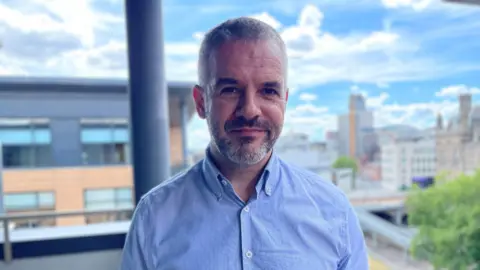 A man with short grey hair and a short grey beard is wearing a blue shirt. He is standing on a balcony with a view of Sheffield city centre behind him