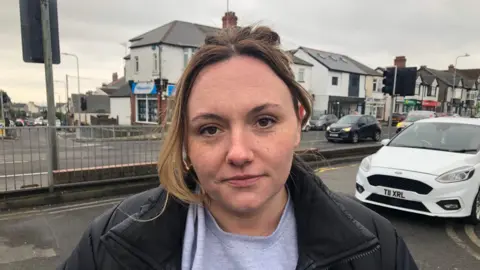 Rachel Williams looking at the camera. She is standing on Newport Road with cars driving behind her. She has brown hair and is wearing a grey jumper and black coat. 