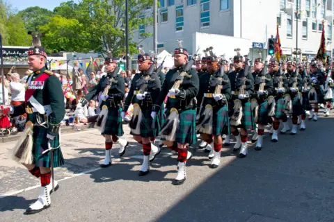 RODDY SIMPSON Soldiers from The Royal Regiment of Scotland marching along a street in full Highland dress, including kilts, and carrying weapons on their shoulders.