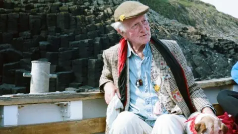 Family handout Peter sits in a boat, which is running along a rocky coastline. He wears a flat cap with a flower pinned to it, a red scarf, a blue shirt and a gingham blazer with lots of badges and patches attached to it. He is stroking a small dog with a red handkerchief tied round its neck. 