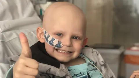 Family Handout A boy in a hospital bed smiling and giving a thumbs up. He has no hair and a plaster with blue teddies securing a drip line to his right cheek. He is wearing a grey dressing gown with stars on it and turquoise pyjamas.