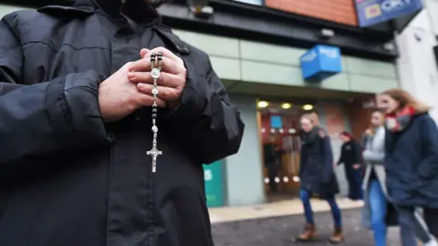 close up shot of the hands of a man holding rosary beads outside an abortion clinic. Female pedestrians are walking out of focus in the background