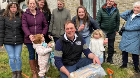 Adults and some children pose with the time capsule which is covered in plastic wrapping 