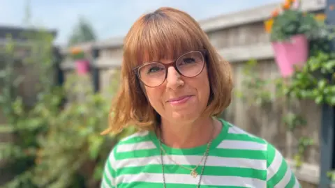 Catherine Warrilow Catherine Warrilow with a light brown bob and fringe, wearing glasses and a green and white horizontal striped t shirt in a garden