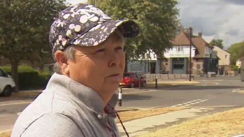 A woman with short grey hair wearing a spotty black and white baseball cap and grey polo shirt. She is standing next to a residential street.
