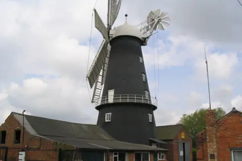 Lincolnshire County Council A black windmill with white sails, and red brick buildings surrounding it on the ground.