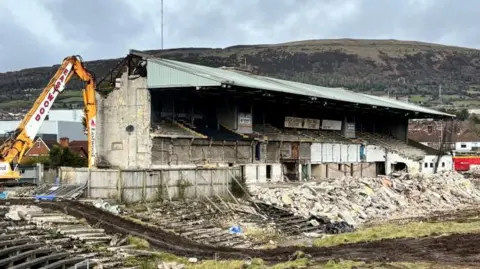 BBC Rubble and bricks are laying on the grass as a yellow digger is knocking down a grandstand. 