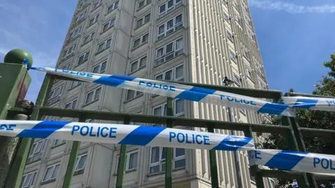 Blue and white police tape tied to a green metal fence at the entrance to the tower block, which is tall and cream.