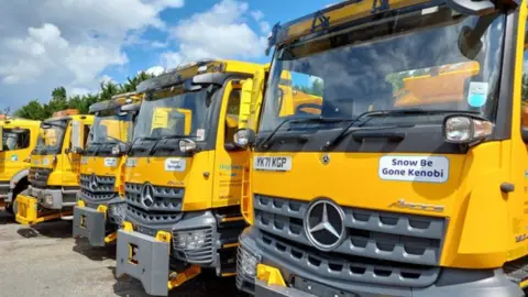 Shropshire Council A line of five yellow gritter lorries, the closest to the camera has a sticker proclaiming 'Snow be Gone Kenobi'