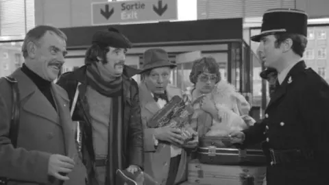 Image from the film Grand Slam, Dewi Pwys Morris second right in the shot in the role of Glyn Lloyd-Evans - dressed in a black long leather coat, scarf and black flat hat, sporting a large moustache. Standing next to him is the actor Windsor Davies, who played the character Mog Jones. Other cast members are present, speaking to a French police officer