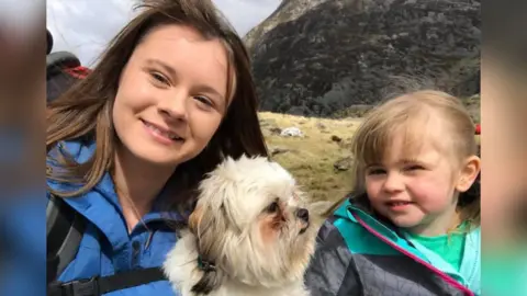 Melonie Bray Melonie Bray smiling at the camera, she has long brown hair and is wearing a blue rain coat. To her right is her daughter Sophie, and their dog. 